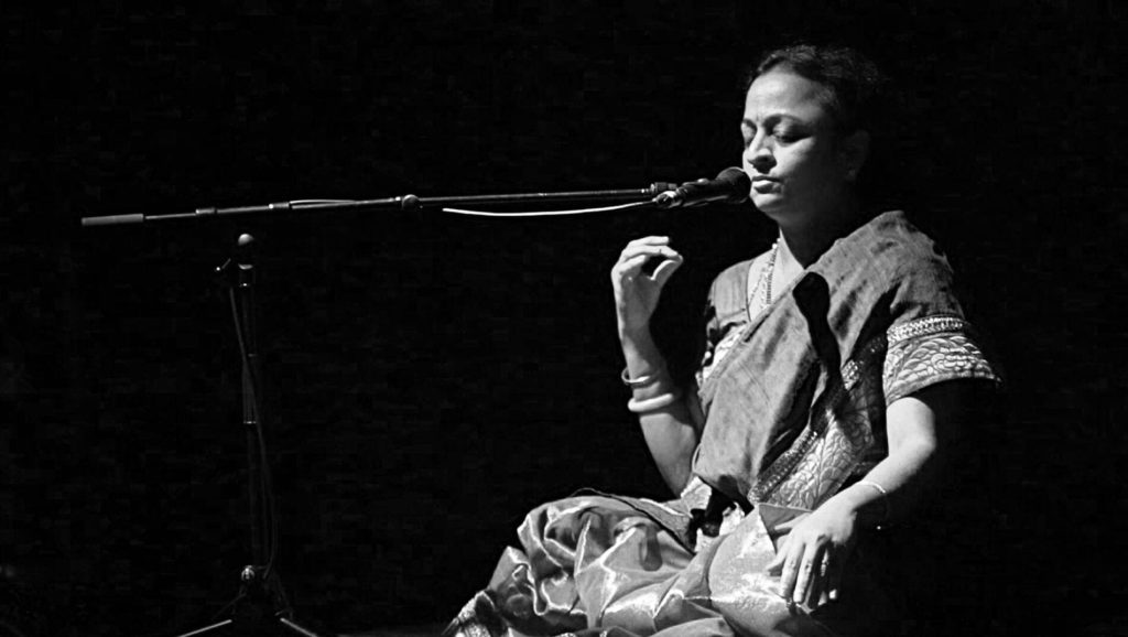 Supriya Nagarajan sitting cross-legged, with her eyes closed and a microphone in front of her mouth