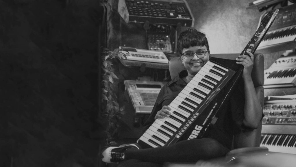 Yshani Perinpanayagam smiling while sitting on a chair and holding a keyboard, with more keyboards in the background