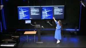 Amelia presenting at the RNCM PGR conference. She is wearing a bright blue dress and is gesturing to a large screen which shows an image of a ballet dancer.