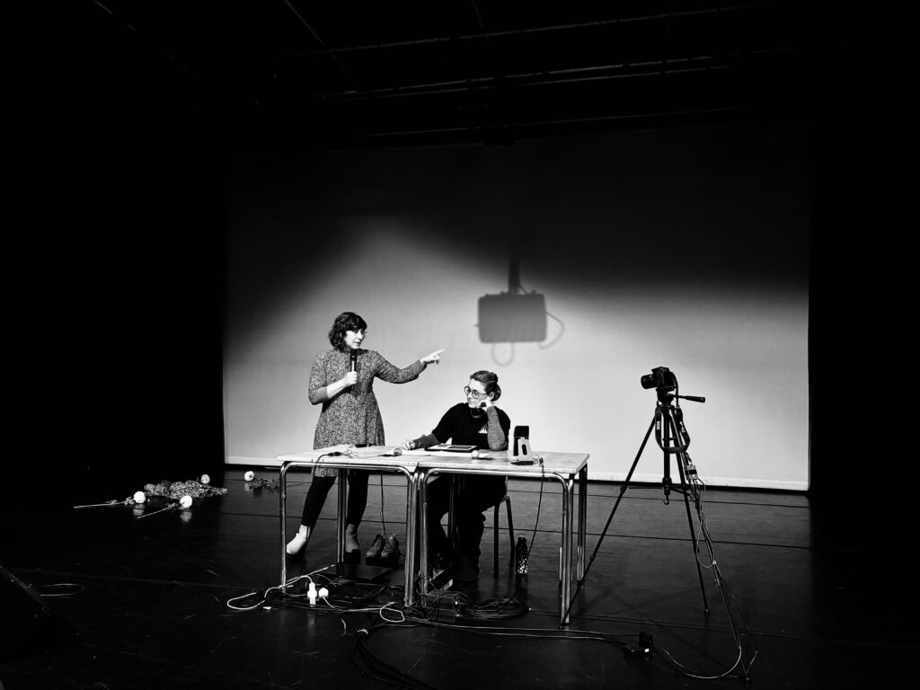 Composer gestures with her left hand. Performer sits at table looking thoughtful.