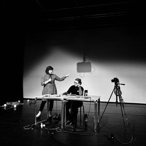 Composer gestures with her left hand. Performer sits at table looking thoughtful.